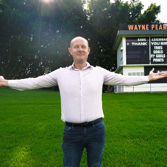 Mayor Darcy Byrne stand in a grassy ovla smiling wiht arms outstretched. Behind him are trees and a sports leader board the reads: 'Wayne Pearce Hill'